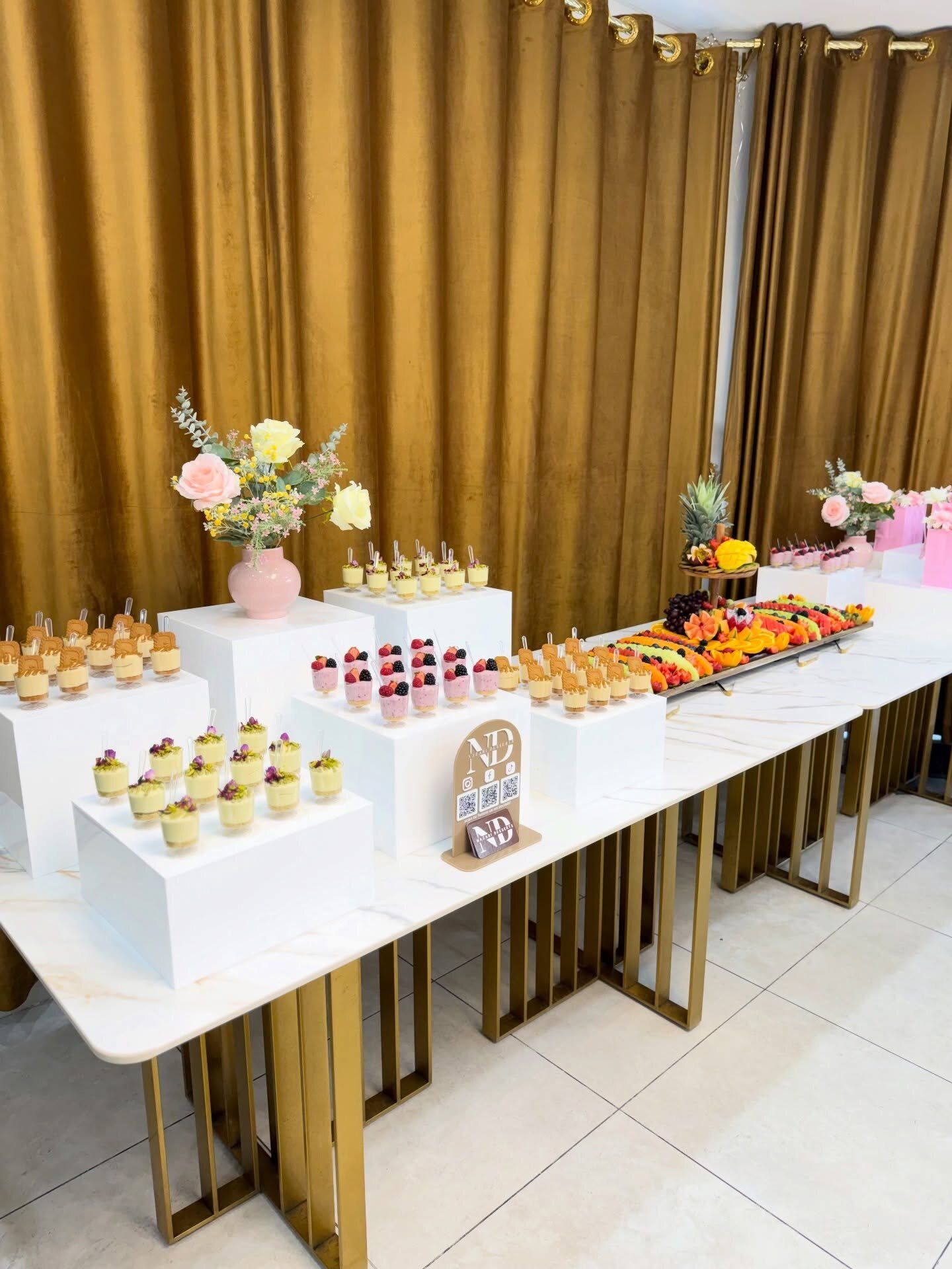 A full event setup with a fruit board, dessert cups and florals in front of gold drapes
