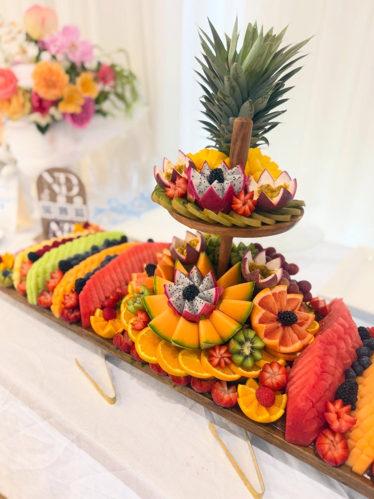 A fruit board with a carved fruit tier, dragon fruit flowers and a pineapple crown, beside fresh florals